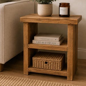 May include: A rectangular wooden side table with a natural finish, featuring a flat top surface and one lower shelf. The top holds a potted green plant and a brown glass candle jar. The lower shelf contains two stacked books and a woven basket.