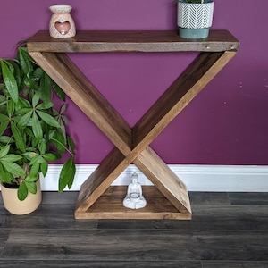 Puede incluir: Una consola de madera con base en forma de X y tapa rectangular. Una peque&ntilde;a estatua de Buda blanca se encuentra en el estante inferior. Una planta en maceta con hojas verdes y una maceta de cer&aacute;mica decorativa est&aacute;n en el estante superior.