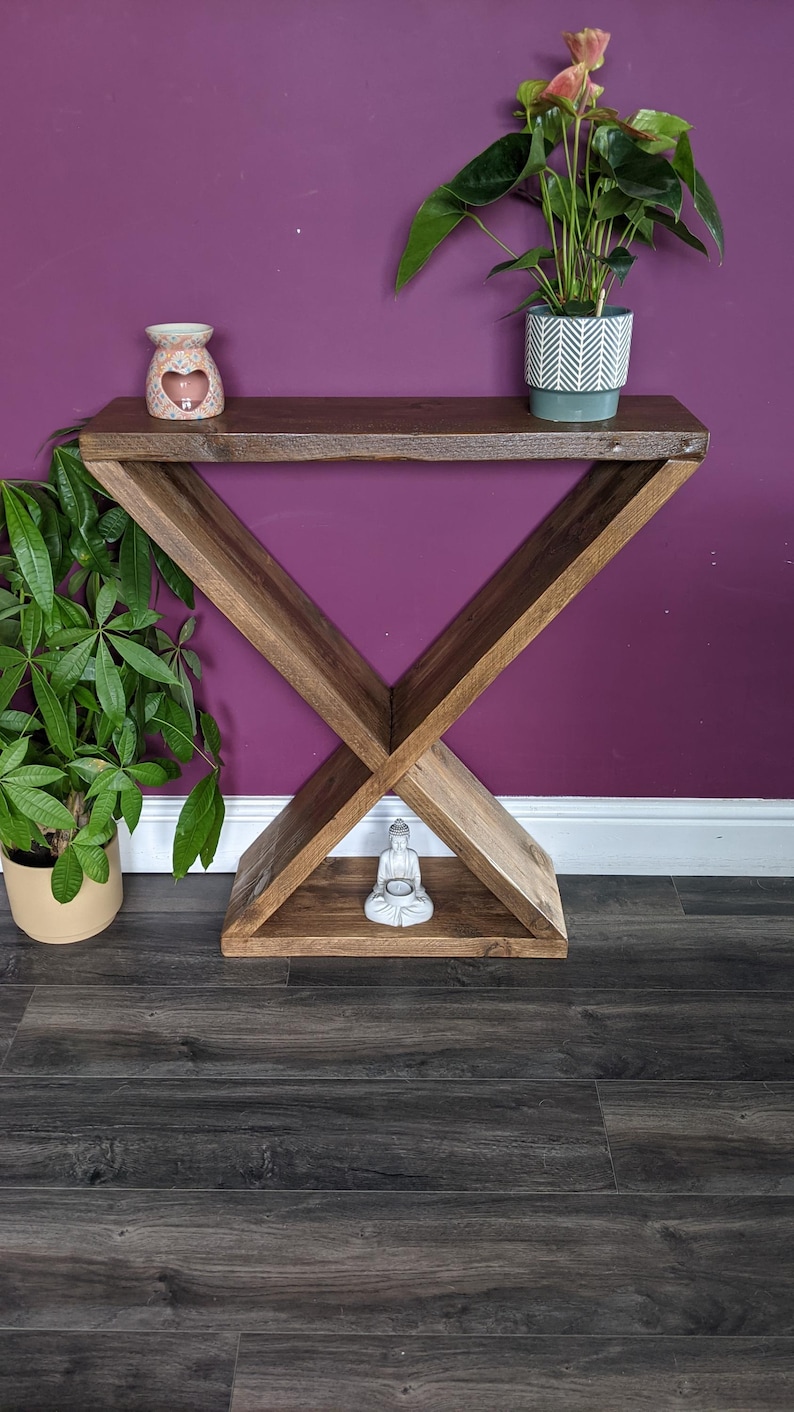 Puede incluir: Una consola de madera con base en forma de X y tapa rectangular. Una peque&ntilde;a estatua de Buda blanca se encuentra en el estante inferior. Una planta en maceta con hojas verdes y una maceta de cer&aacute;mica decorativa est&aacute;n en el estante superior.