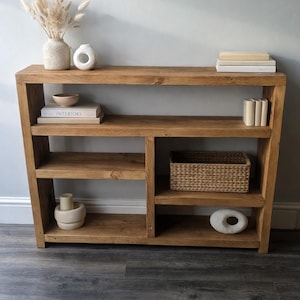 May include: A wooden bookcase with a natural finish, featuring multiple shelves and compartments. It's styled with decorative items including books, vases, and a woven basket. The unit is designed for storage and display, adding a touch of rustic charm.