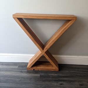 Puede incluir: Una consola de madera con un dise&ntilde;o geom&eacute;trico &uacute;nico. La mesa est&aacute; construida con madera de color marr&oacute;n claro y presenta una base en forma de X que sostiene una parte superior rectangular. La mesa mide aproximadamente 76 cm de largo y 30 cm de ancho.