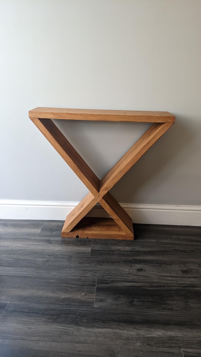 Puede incluir: Una consola de madera con un dise&ntilde;o geom&eacute;trico &uacute;nico. La mesa est&aacute; construida con madera de color marr&oacute;n claro y presenta una base en forma de X que sostiene una parte superior rectangular. La mesa mide aproximadamente 76 cm de largo y 30 cm de ancho.