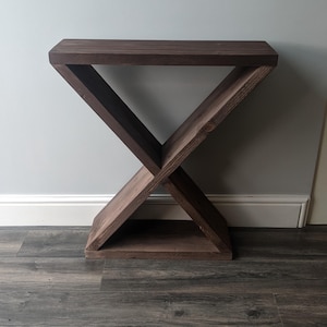 Puede incluir: Una mesa auxiliar de madera marr&oacute;n oscuro con una base en forma de X y una parte superior rectangular. La mesa est&aacute; hecha de madera maciza y tiene un acabado natural. El dise&ntilde;o es moderno y minimalista, adecuado para una sala de estar o dormitorio.