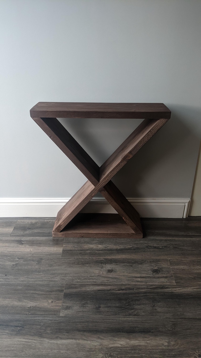 Puede incluir: Una mesa auxiliar de madera marr&oacute;n oscuro con una base en forma de X y una parte superior rectangular. La mesa est&aacute; hecha de madera maciza y tiene un acabado natural. El dise&ntilde;o es moderno y minimalista, adecuado para una sala de estar o dormitorio.