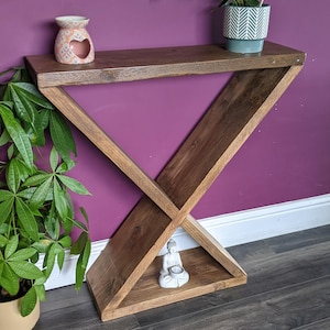 Puede incluir: Una consola de madera oscura con un dise&ntilde;o geom&eacute;trico &uacute;nico. La mesa marr&oacute;n oscuro presenta una base triangular y una parte superior rectangular. Un peque&ntilde;o difusor de aceite de cer&aacute;mica y una planta en maceta est&aacute;n en el estante superior. Una peque&ntilde;a estatua de Buda est&aacute; en el estante inferior.