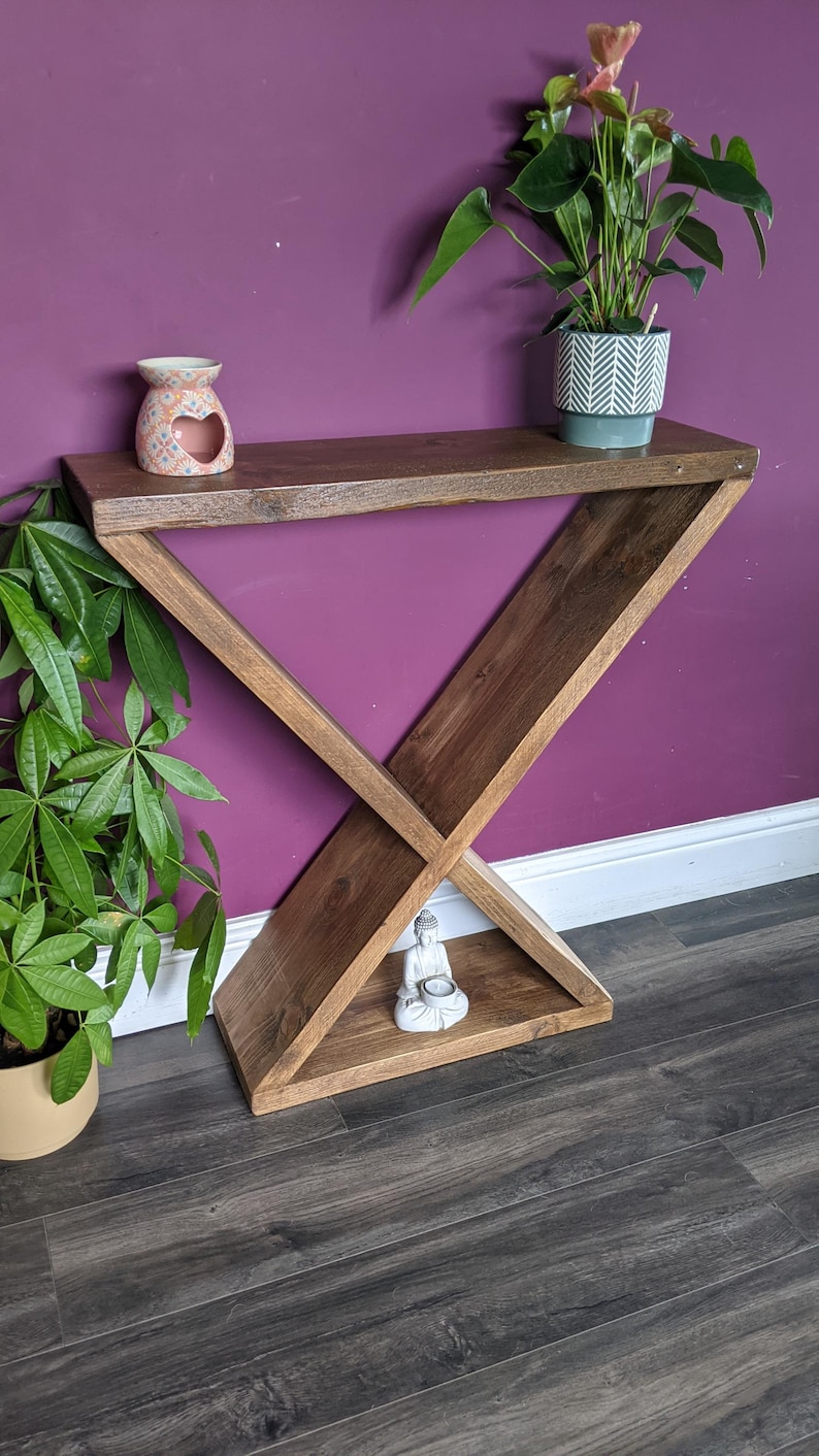 Puede incluir: Una consola de madera oscura con un dise&ntilde;o geom&eacute;trico &uacute;nico. La mesa marr&oacute;n oscuro presenta una base triangular y una parte superior rectangular. Un peque&ntilde;o difusor de aceite de cer&aacute;mica y una planta en maceta est&aacute;n en el estante superior. Una peque&ntilde;a estatua de Buda est&aacute; en el estante inferior.