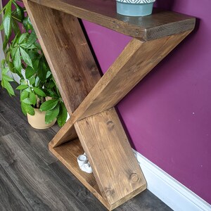 Puede incluir: Una consola de madera hecha a mano con un dise&ntilde;o &uacute;nico en forma de X. La mesa est&aacute; te&ntilde;ida de un marr&oacute;n c&aacute;lido y tiene un estante. Un peque&ntilde;o jarr&oacute;n decorativo y una planta en maceta est&aacute;n encima. La mesa mide 122 cm de largo.