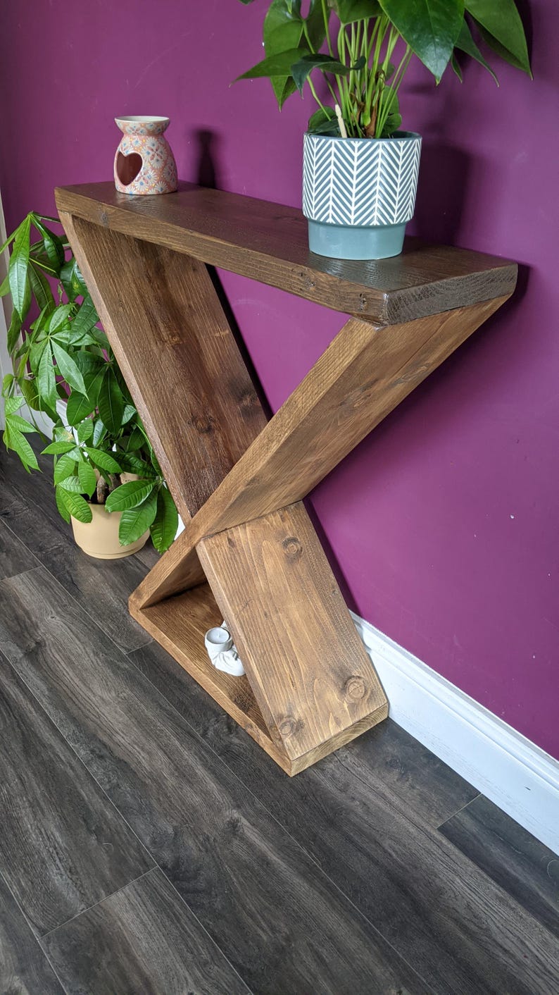 Puede incluir: Una consola de madera hecha a mano con un dise&ntilde;o &uacute;nico en forma de X. La mesa est&aacute; te&ntilde;ida de un marr&oacute;n c&aacute;lido y tiene un estante. Un peque&ntilde;o jarr&oacute;n decorativo y una planta en maceta est&aacute;n encima. La mesa mide 122 cm de largo.