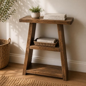 May include: A brown wooden console table with a trapezoidal frame, featuring a top surface, a middle shelf, and a bottom shelf. The top holds a small potted plant and two books titled The Kinfolk Home and Live Beautifully. A woven basket with a book titled This Is Home sits on the middle shelf.