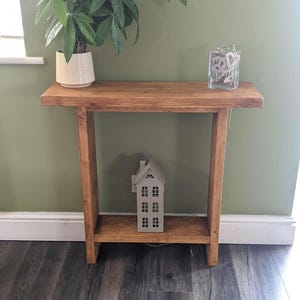 May include: A rectangular wooden console table with a natural brown finish, featuring a flat top surface and a lower shelf. A potted green plant sits on the left side of the top, while a small grey house-shaped lantern rests on the bottom shelf.