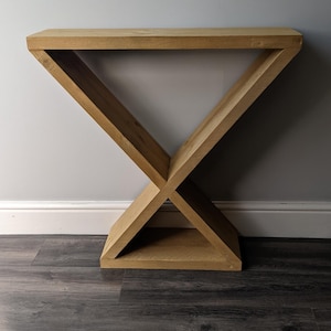 Puede incluir: Una consola de madera con una base en forma de X. La mesa est&aacute; hecha de madera clara y tiene una parte superior rectangular. La base est&aacute; formada por dos vigas que se cruzan, creando un dise&ntilde;o geom&eacute;trico. La mesa mide 76 cm de largo.