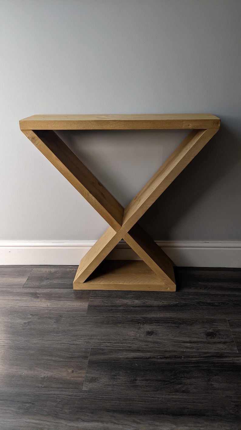 Puede incluir: Una consola de madera con una base en forma de X. La mesa est&aacute; hecha de madera clara y tiene una parte superior rectangular. La base est&aacute; formada por dos vigas que se cruzan, creando un dise&ntilde;o geom&eacute;trico. La mesa mide 76 cm de largo.