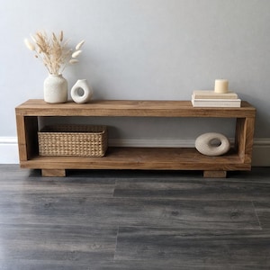 May include: A rectangular, wooden coffee table with a natural finish. The table features a lower shelf with a woven basket and a decorative stone. On the top shelf are books, a candle, and two ceramic vases with dried plants. The table is 122 cm long.