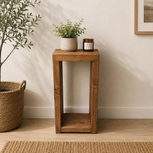 May include: A tall, rectangular wooden side table with a medium brown finish and a visible wood grain. The table features a flat top surface, two vertical side supports, and a bottom shelf. A small potted green plant and a brown candle sit on top.