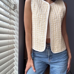 May include: A sleeveless, quilted vest in a light yellow and white woven pattern. The vest has a V-neck and is trimmed with white binding. The person is wearing blue jeans.