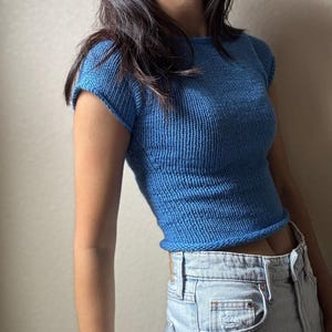 May include: A short-sleeved, cropped blue knit top. The top has a ribbed texture and a rounded neckline. The garment is paired with light-wash denim jeans. The overall style is casual and modern.