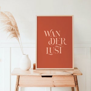 Puede incluir: Una impresión enmarcada con el texto "Wanderlust" en rosa claro sobre un fondo naranja quemado. La impresión está sobre una mesa de madera con un jarrón blanco y una cesta de mimbre.