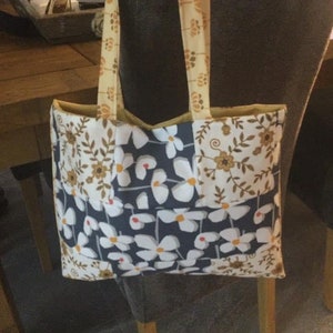 Puede incluir: Una bolsa de mano con un dise&ntilde;o de patchwork que presenta flores blancas sobre un fondo azul oscuro, flores blancas sobre un fondo crema y patrones florales marrones sobre un fondo crema. La bolsa tiene un forro de tela amarillo y correas amarillas.