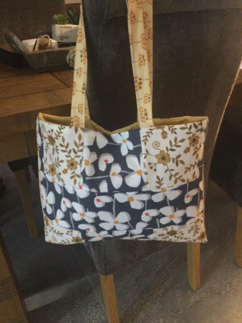 Puede incluir: Una bolsa de mano con un dise&ntilde;o de patchwork que presenta flores blancas sobre un fondo azul oscuro, flores blancas sobre un fondo crema y patrones florales marrones sobre un fondo crema. La bolsa tiene un forro de tela amarillo y correas amarillas.