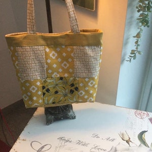 Puede incluir: Una bolsa de mano de lona amarilla y blanca con un estampado floral en uno de los cuadrados. La bolsa tiene dos asas largas y est&aacute; sentada sobre una mesa blanca con un dise&ntilde;o floral.