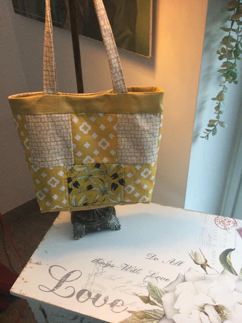 Puede incluir: Una bolsa de mano de lona amarilla y blanca con un estampado floral en uno de los cuadrados. La bolsa tiene dos asas largas y est&aacute; sentada sobre una mesa blanca con un dise&ntilde;o floral.