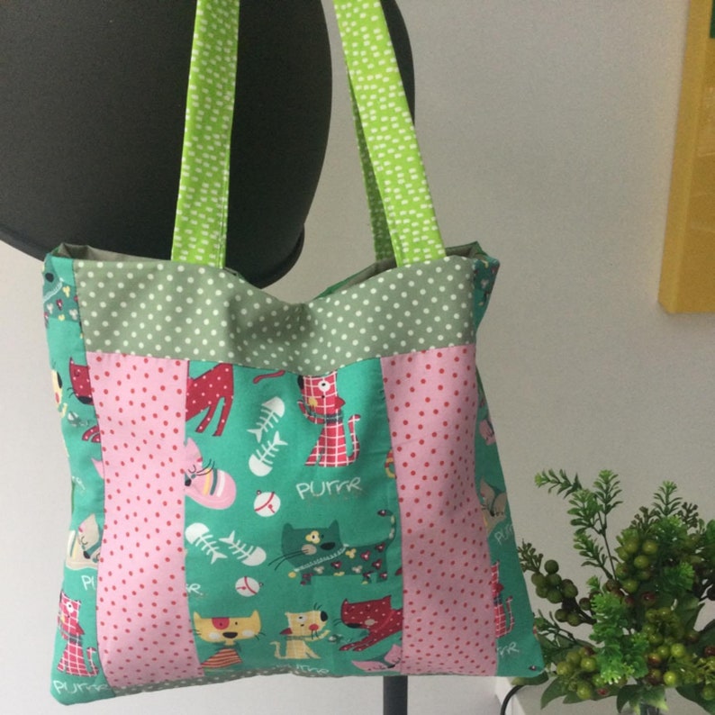 Op de afbeelding: Een groene, roze en witte tote bag met een kattenprint. De tas heeft een groene stof met witte stippen aan de boven- en onderkant, en een roze stof met witte stippen in het midden. De kattenprint is op een groene achtergrond en toont katten met visgraten, honkbalballen en het woord "Purrr".