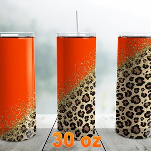 Pode incluir: Três canecas de aço inoxidável de 887 ml com tampas e canudos. As canecas têm um fundo laranja com glitter dourado e um design de estampa de leopardo. O texto "30 oz" é visível na parte inferior.