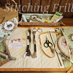 May include: A quilted cream-colored embroidery organizer with a floral and bee pattern. The organizer holds embroidery floss, scissors, needles, and a wooden hoop. The text "Stitching Frills" is visible on a business card.