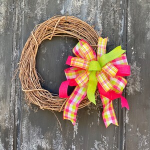 May include: A grapevine wreath adorned with a vibrant bow. The bow features a plaid pattern in shades of pink, yellow, and white, complemented by solid pink and lime green ribbons. This decorative wreath is perfect for adding a touch of color to any door or wall.