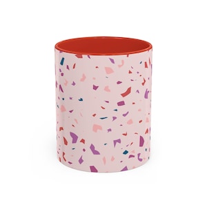 May include: A ceramic coffee mug with a light pink exterior adorned with a terrazzo pattern in shades of pink, purple, red, and teal. The interior of the mug is a vibrant red color.
