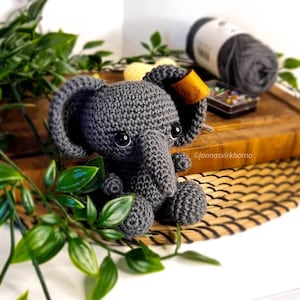 May include: A gray crocheted elephant toy with black eyes and a brown leather tag that says "jonnasvirkhorna". The elephant is sitting on a woven mat.