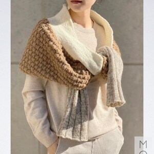 Può includere: Una sciarpa di lana marrone, bianca e beige con un motivo testurizzato. La sciarpa è indossata intorno al collo di una persona che indossa un maglione e pantaloni beige. Il testo "MONM Knitting" è visibile nell'angolo in basso a destra dell'immagine.