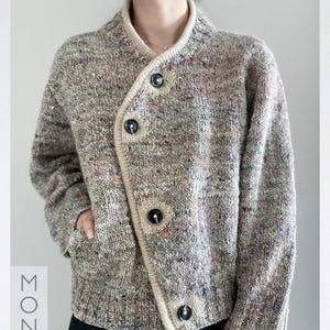 Puede incluir: Cárdigan de punto jaspeado en tonos marrón, beige y motas de color. El suéter presenta un cuello chal, tres botones negros grandes y un bolsillo. El ribete es de color beige claro. La palabra "Knitting" es visible en la esquina inferior izquierda.