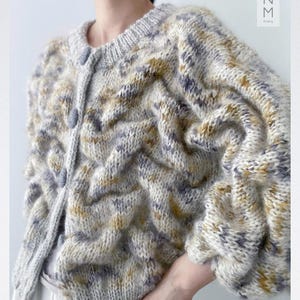May include: A chunky knit cardigan featuring a textured, wavy pattern in cream, mustard yellow, and grey yarn. The garment has a round neckline, long sleeves with gathered cuffs, and a front button closure with matching grey buttons. The text MOZM is visible.