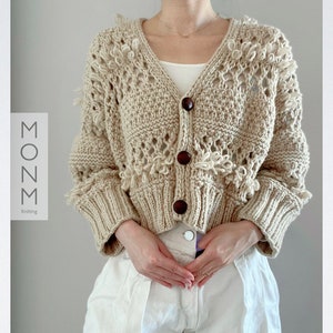 May include: A beige, open-weave cardigan with a V-neck and three large, dark brown buttons. The sleeves and bottom hem are ribbed. The cardigan has a textured, fringed design. The text "MONN Knitting" is on the left side.