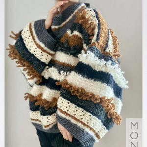 Puede incluir: Un suéter a rayas con hilo marrón, blanco, negro y gris. El suéter tiene cuello alto y mangas largas con detalles de flecos. El suéter está tejido con un punto texturizado.