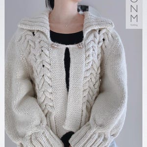 May include: A cream-colored knitted cardigan with a cable knit pattern and a large collar. The cardigan has two buttons on the front and long sleeves.