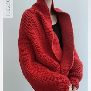 Könnte beinhalten: Ein leuchtend roter Strickcardigan mit breitem Schalkragen. Das grob gestrickte Kleidungsstück hat eine lockere Passform und lange Ärmel. Der Text "MOZM Knitting" ist vertikal auf der linken Seite aufgedruckt.