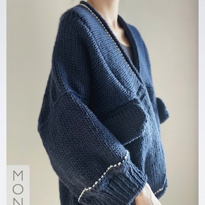 Pode incluir: Um cardigan de malha azul marinho com decote em V e acabamento branco ao redor do pescoço e punhos. O cardigan é feito de um fio macio e grosso e tem um caimento relaxado.