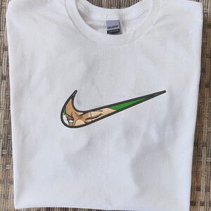 Op de afbeelding: Wit T-shirt met een groen en zwart geborduurd ontwerp dat lijkt op een populair merklogo, maar met het gezicht van een stripfiguur erin verwerkt. Het shirt is gemaakt van een zacht materiaal en heeft een ronde hals.