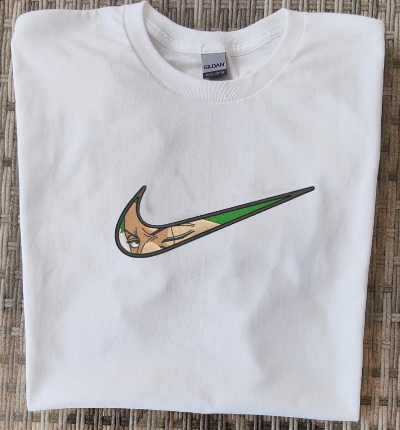 Op de afbeelding: Wit T-shirt met een groen en zwart geborduurd ontwerp dat lijkt op een populair merklogo, maar met het gezicht van een stripfiguur erin verwerkt. Het shirt is gemaakt van een zacht materiaal en heeft een ronde hals.