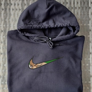 Op de afbeelding: Een zwarte hoodie met een capuchon met trekkoord. Het Nike swoosh-logo is aangepast met een cartoonkarakterontwerp in tinten bruin, groen en wit.