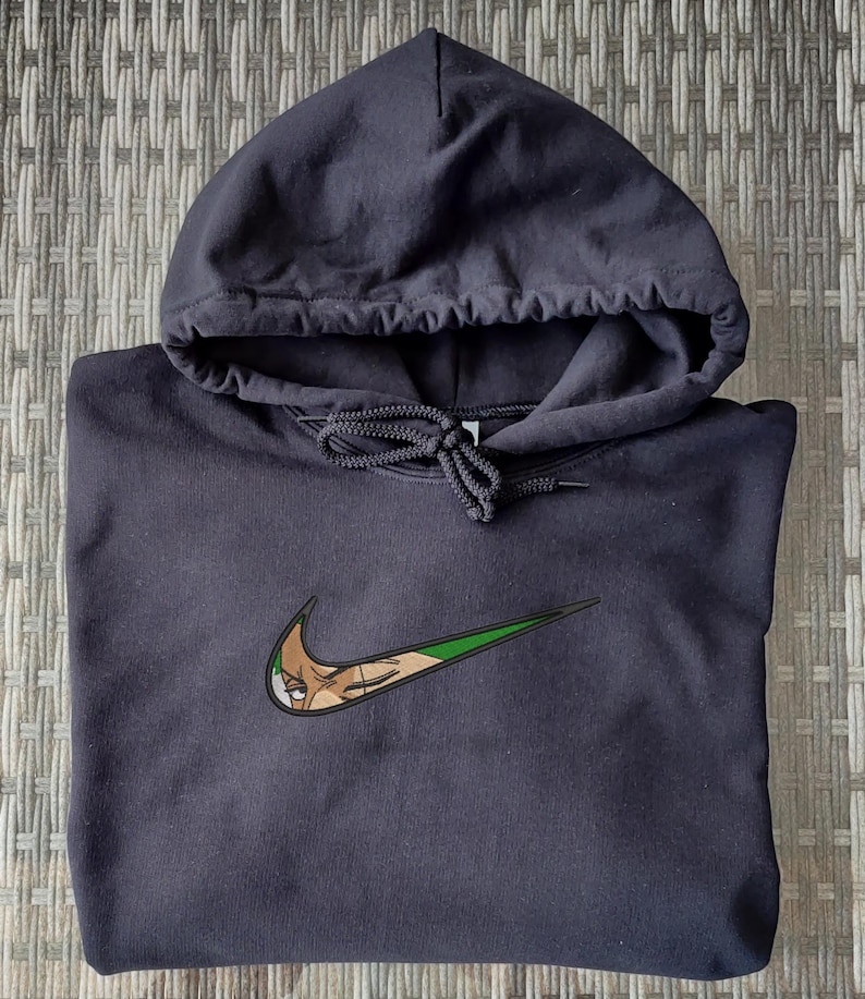 Op de afbeelding: Een zwarte hoodie met een capuchon met trekkoord. Het Nike swoosh-logo is aangepast met een cartoonkarakterontwerp in tinten bruin, groen en wit.