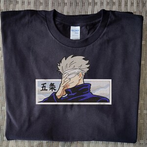 May include: Black t-shirt with an embroidered image of a character with a blindfold, wearing a blue jacket and a white bandage over their eyes. The character is holding their hand over their eyes. The text "五条" is embroidered in white on a white background.