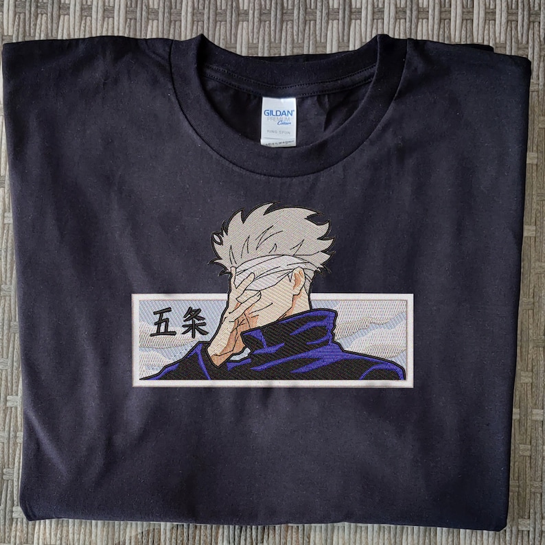 May include: Black t-shirt with an embroidered image of a character with a blindfold, wearing a blue jacket and a white bandage over their eyes. The character is holding their hand over their eyes. The text "五条" is embroidered in white on a white background.
