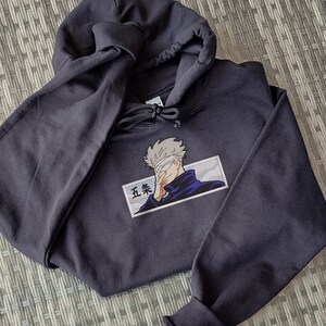 May include: A black hooded sweatshirt with a white and blue embroidered image of a character with a blindfold and the text "五条" on the front.