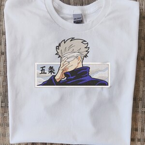 May include: White t-shirt with an embroidered patch featuring a character with a blindfold, a blue jacket, and the text "五条" in Japanese.