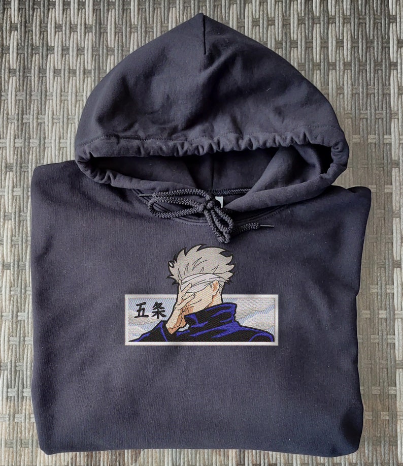 May include: Black hooded sweatshirt with an embroidered image of a character with a blindfold, wearing a blue shirt and a white headband. The character is holding their hand to their face. The text "五条" is embroidered below the character.