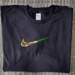 Op de afbeelding: Zwart t-shirt met een geborduurd ontwerp in groen en beige, dat lijkt op een Nike swoosh, met het gezicht van een personage. Het t-shirt is gemaakt van een zacht materiaal, geschikt voor casual wear.