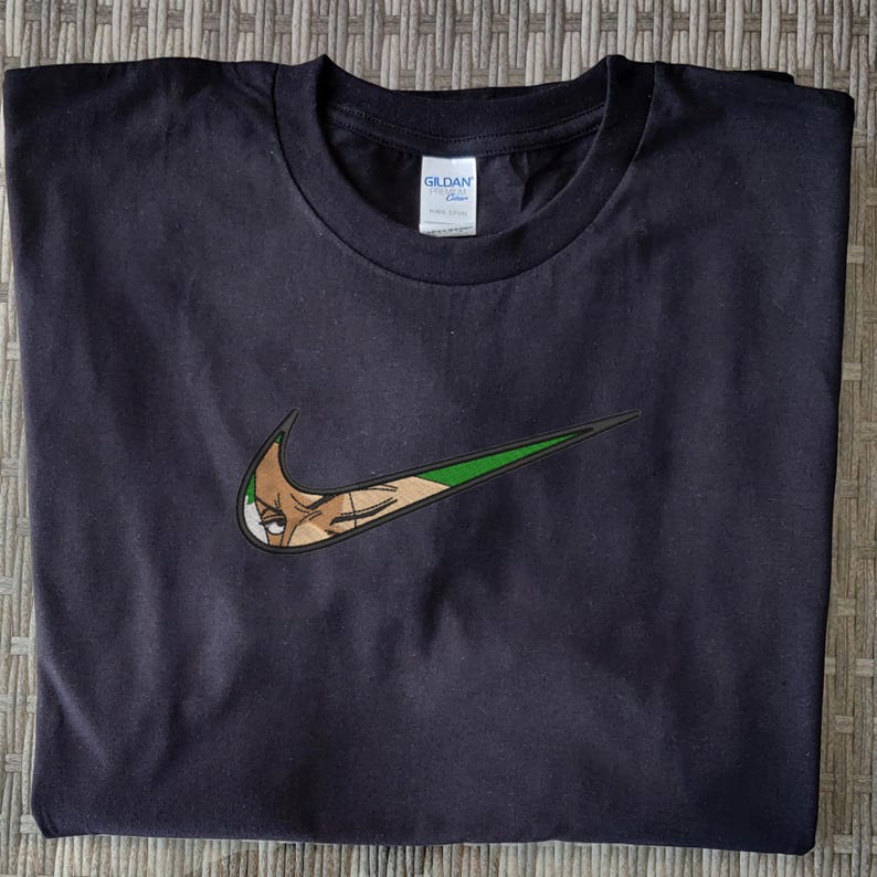 Op de afbeelding: Zwart t-shirt met een geborduurd ontwerp in groen en beige, dat lijkt op een Nike swoosh, met het gezicht van een personage. Het t-shirt is gemaakt van een zacht materiaal, geschikt voor casual wear.